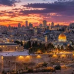 הטפט מציג עיר בשקיעה עם כיפה זהובה, שמיים כתומים וסגולים ואורות עירוניים חמימים.