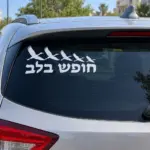 מדבקה עם המילים 'חופש בלב' וצלליות ציפורים על השמשה האחורית של רכב בחניון עירוני.