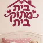 מדבקה עם טקסט עברי, בית ולבבות על קיר בחדר ילדים עם צעצועים וארון צבעוני.