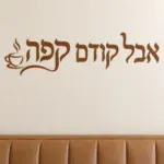 מדבקת טקסט עברי עם כוס קפה שחורה על קיר בסלון עם ספה אפורה ושולחן עץ כהה.
